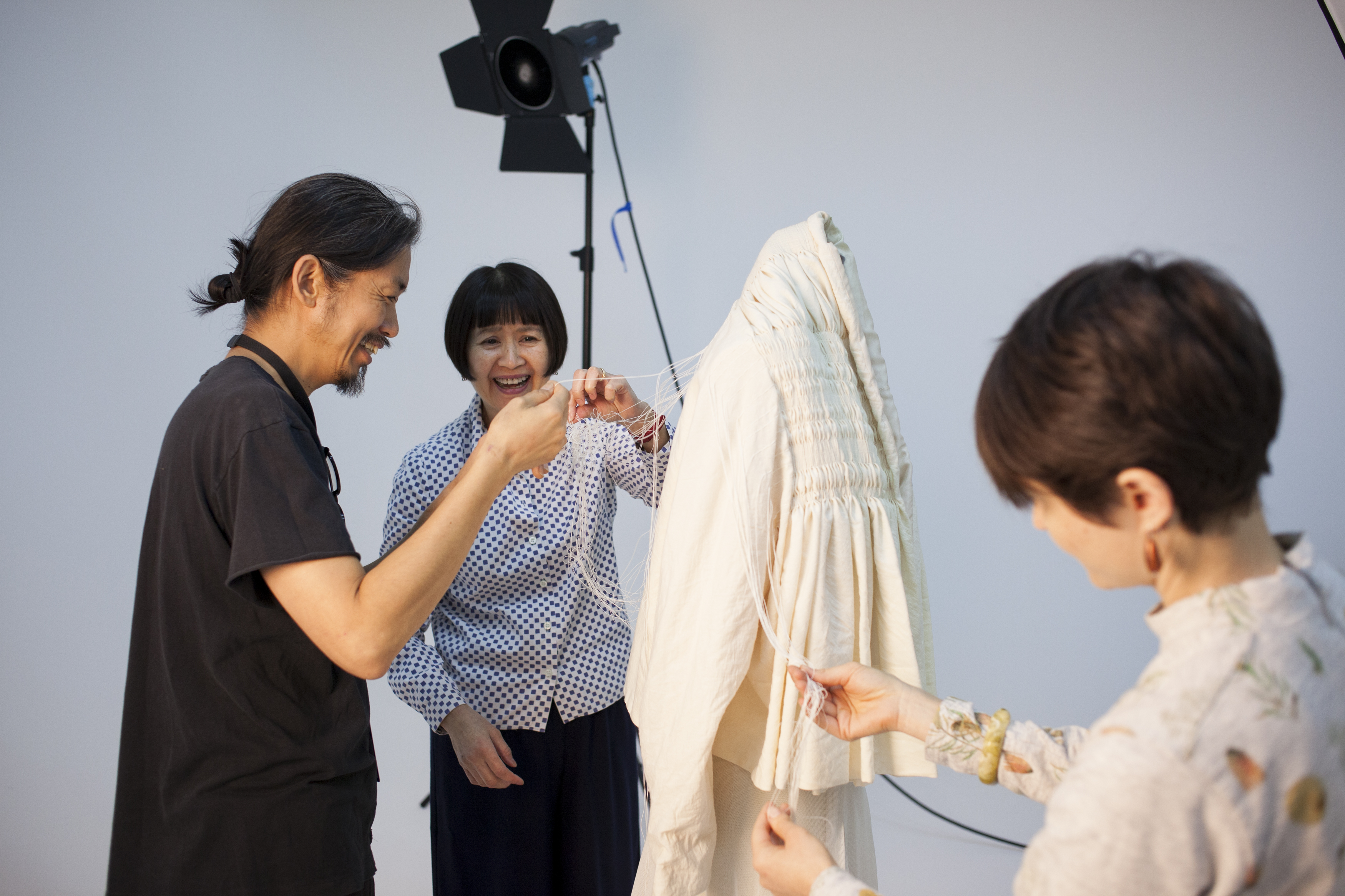 A man and a woman untangling long, linen threads on the back of a woollen jacket while another woman untangles threads on the front of the jacket. There is a stand with camera equipment mounted on it in the background.